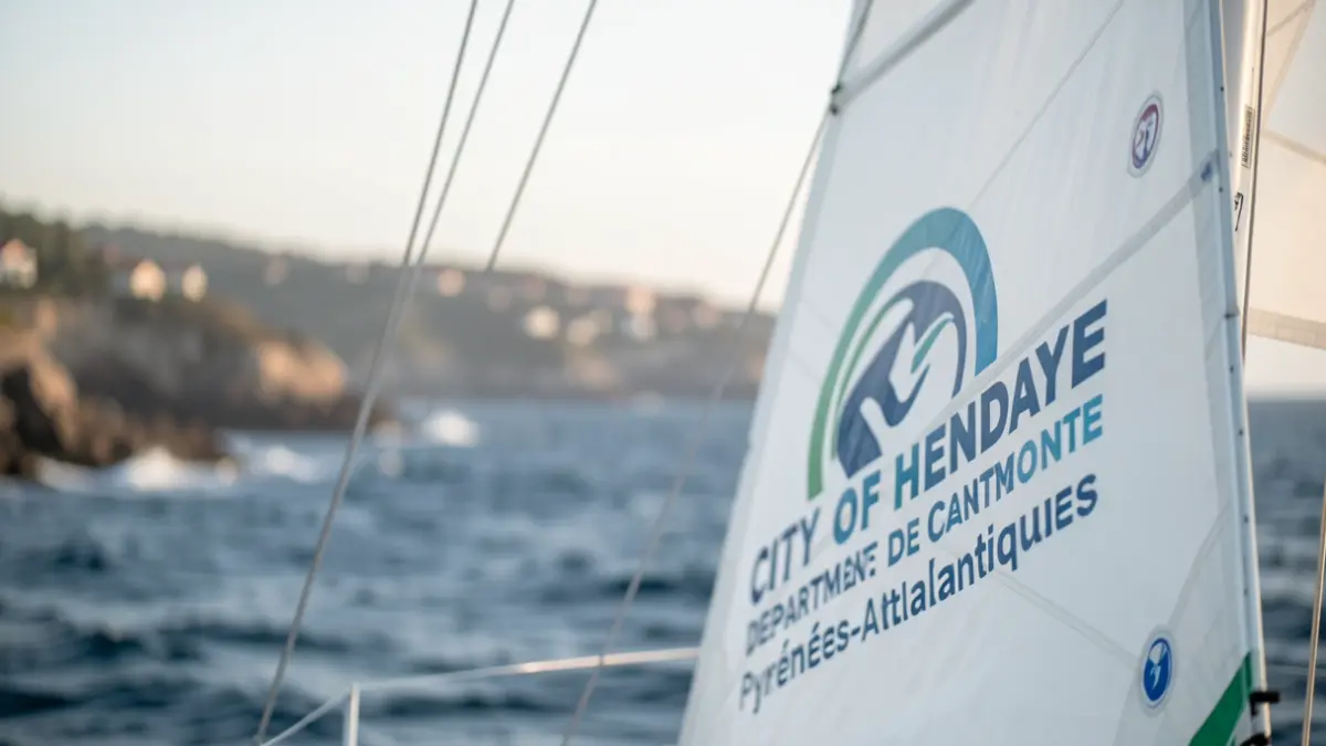 Voilier de course avec les logos de la Ville d'Hendaye et du Département des Pyrénées-Atlantiques, en mer.